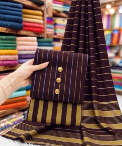 An indoor display of unstitched 3-piece suit PR-142, highlighting its unique check-styled shirt, trouser, and dupatta on a traditional braided cot (charpai) with a vintage handloom backdrop.